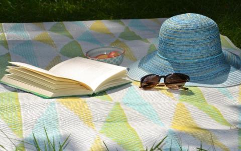 Picknickdecke mit aufgeschlagenem Buch, Sonnenhut und Sonnenbrille drauf